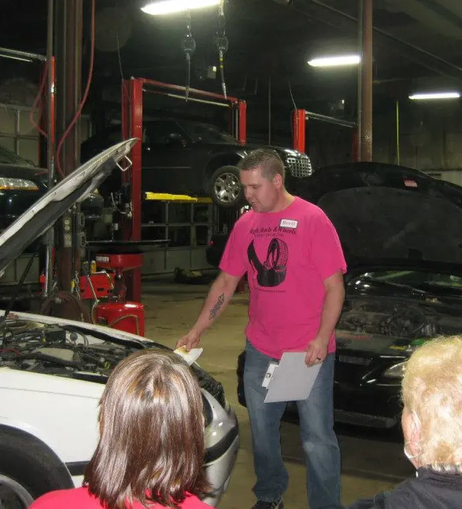 Auto repair technician leading a workshop in a garage, holding papers and pointing at a car engine while explaining maintenance steps to a small group.